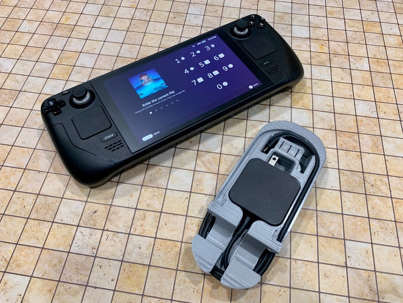May include: A black handheld gaming device with a touchscreen display showing a number pad and the text "Enter the current PIN". The device is lying on a beige tiled surface next to a gray 3D printed charging stand with a black power adapter.