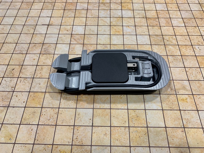 May include: A 3D printed gray plastic holder for a phone charger with a black charging pad and a black cord.