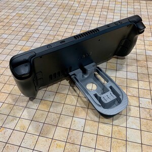 May include: A black handheld gaming console with a gray 3D printed stand. The console has a textured back and a small screen. The stand is designed to hold the console upright.