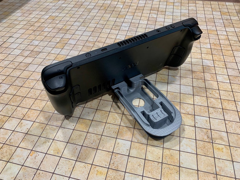 May include: A black handheld gaming console with a gray 3D printed stand. The console has a textured back and a small screen. The stand is designed to hold the console upright.