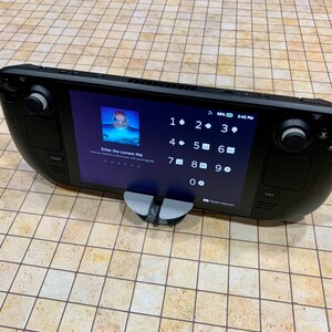 May include: A black handheld gaming device with a touchscreen display showing a numeric keypad and the text "Enter the current PIN". The device is resting on a gray stand.