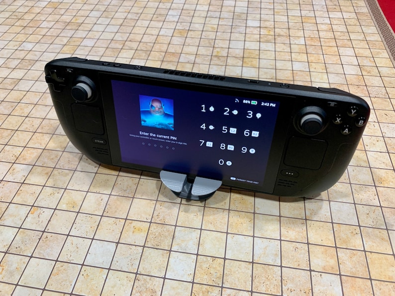 May include: A black handheld gaming device with a touchscreen display showing a numeric keypad and the text "Enter the current PIN". The device is resting on a gray stand.