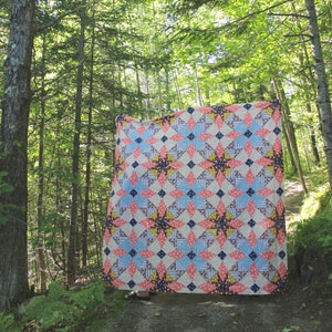 Könnte beinhalten: Eine große Patchwork-Decke mit einem geometrischen Muster in Rosa-, Blau- und Brauntönen. Die Decke hängt in einer Waldlandschaft.