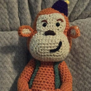 May include: Handmade crochet monkey doll in orange, cream, and purple. The monkey has a smiling face, a small hat, and green straps. The doll is made of yarn and has a tail.