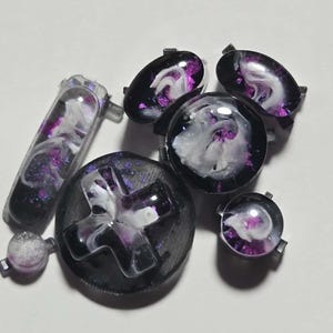 May include: A selection of black and purple resin gaming controller buttons and accessories. The buttons feature a swirling white and purple design. The collection includes a D-pad, a start button, and other buttons.