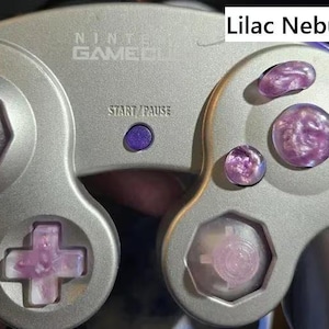 Może przedstawiać: Srebrny kontroler Nintendo GameCube z wzorem "Lilac Nebula Set". Kontroler ma guziki i drążki w kolorze liliowym. Przycisk Start/Pause jest fioletowy. Kontroler ma klasyczny design.