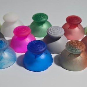 Może przedstawiać: Zestaw 18 kolorowych zamiennych gałek analogowych do kontrolerów gier wideo. Gałki analogowe są wykonane z gładkiego, błyszczącego plastiku i dostępne są w różnych kolorach, w tym czerwonym, niebieskim, zielonym, różowym, białym i czarnym.