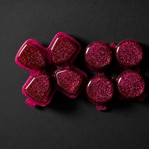 May include: A set of replacement gaming controller buttons in a vibrant magenta colour with a dense, sparkling glitter finish. The set includes a four-way directional pad and four individual circular action buttons, all featuring a glossy surface.