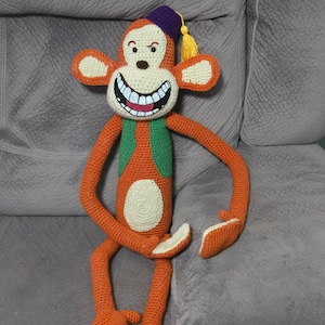 May include: Crocheted monkey doll in orange, white, green, and purple. The monkey has a large, smiling mouth, a purple fez with a yellow tassel, and long arms and legs. The doll is sitting on a gray couch.
