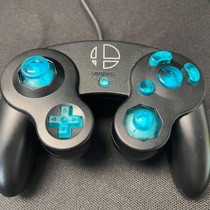 May include: Black video game controller with blue buttons and joysticks. The controller features the Super Smash Bros. logo and a start/pause button. Designed for gaming.