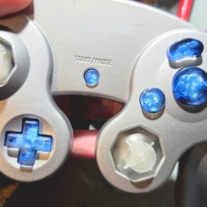 Peut inclure: Manette de jeu vidéo argentée avec des boutons translucides bleus et un pavé directionnel. La manette porte la mention "START/PAUSE" et un joystick hexagonal transparent. Les boutons ont un design bleu nuageux.