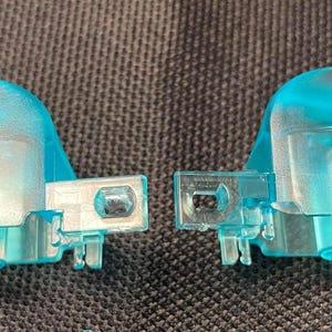 Può includere: Due pulsanti trasparenti azzurri per controller. I pulsanti hanno una parte superiore arrotondata e una base rettangolare con un connettore. Il pulsante sinistro ha la lettera "L" stampata.