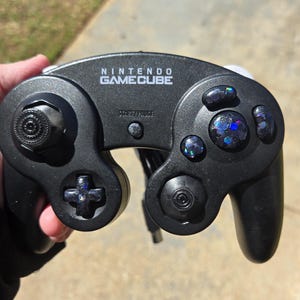 May include: Black Nintendo GameCube controller with the brand name printed on the top. The controller has a directional pad, analog stick, and buttons. The buttons have a glittery, iridescent design.