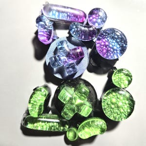 May include: A collection of colorful, translucent ear gauges in various shapes. The gauges feature shades of purple, blue, and green, with some displaying a speckled or cross design. The items are arranged on a light gray surface.