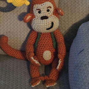 May include: A handmade crochet monkey doll in shades of orange, cream, and green. The monkey has a cream-colored face, a black smile, and a purple hat with a yellow tassel. The doll is approximately 12 inches tall.