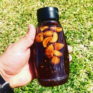 Puede incluir: Una botella de vidrio transparente llena de un líquido marrón oscuro y que contiene dientes de ajo enteros. La botella tiene una tapa negra y una superficie texturizada.