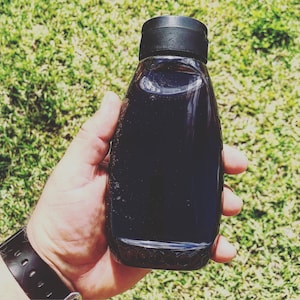 Könnte beinhalten: Eine schwarze Glasflasche mit schwarzem Plastikverschluss. Die Flasche ist mit einer dunklen Flüssigkeit gefüllt.