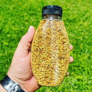 Op de afbeelding: Een doorzichtige plastic fles gevuld met gele stuifmeelkorrels. De fles heeft een zwarte dop en wordt in de hand van een persoon gehouden.