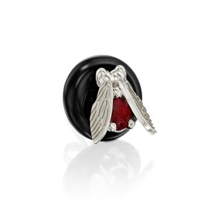Pode incluir: Um amuleto de prata com uma pedra preciosa vermelha e um design de borboleta detalhado. O amuleto está preso a uma conta preta.