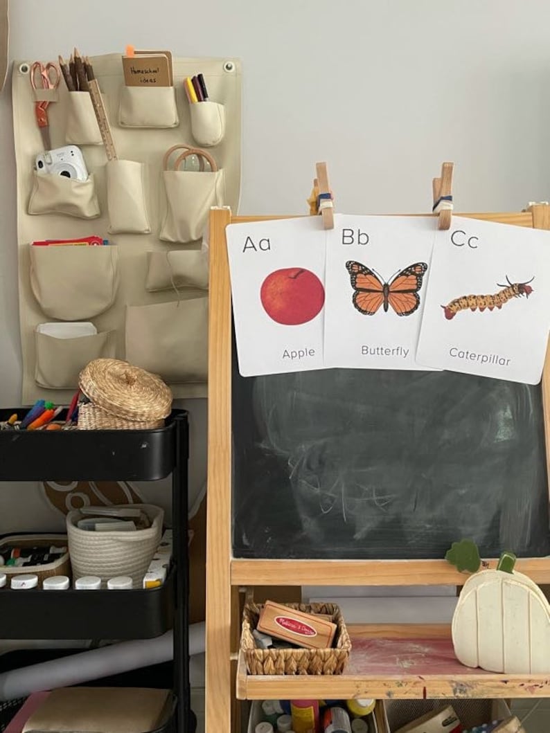 May include: A wooden easel with a chalkboard and three alphabet flashcards. The flashcards show the letters A, B, and C with pictures of an apple, butterfly, and caterpillar. The easel has a shelf with a small wooden box and a white pumpkin.