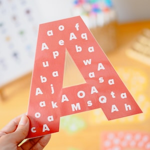 Puede incluir: Una letra A grande de color rojo, troquelada en cartulina, con un patrón de varias letras mayúsculas y minúsculas blancas impresas en su superficie. La letra se sostiene en alto, mostrando un acabado mate plano con bordes limpios.
