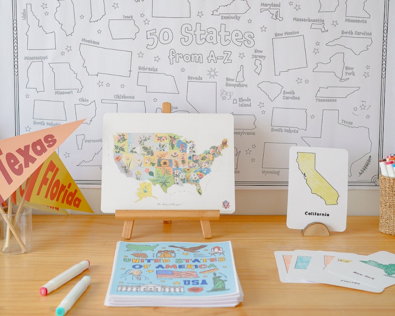 Pu&ograve; includere: Un cavalletto di legno che mostra una mappa colorata degli Stati Uniti, con ogni stato illustrato con un fiore diverso. Un set di pennarelli colorati e una pila di libri intitolati "United States of America" sono su un tavolo di legno. Un piccolo cartoncino con una mappa della California &egrave; su un supporto di legno.
