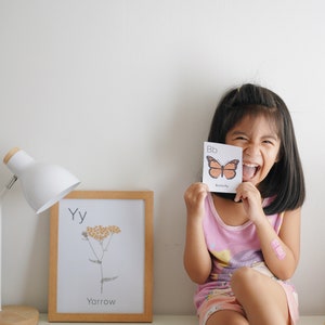May include: A young girl is holding up a flashcard with the letter "B" and a picture of a butterfly. The card says "Bb Butterfly". The girl is smiling and laughing. There are other flashcards on the table, including one with the letter "Y" and a picture of a yarrow plant.