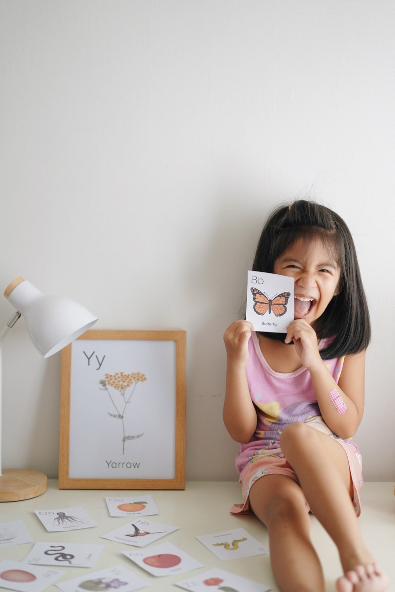 May include: A young girl is holding up a flashcard with the letter "B" and a picture of a butterfly. The card says "Bb Butterfly". The girl is smiling and laughing. There are other flashcards on the table, including one with the letter "Y" and a picture of a yarrow plant.