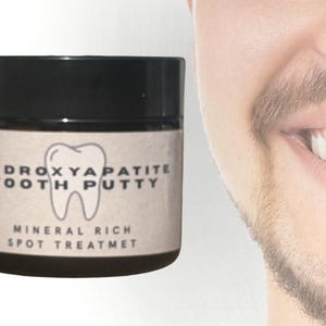 May include: A jar of Hydroxyapatite Tooth Putty with a black lid. The label features a tooth graphic and text that reads "Mineral Rich Spot Treatment". A smiling person is visible in the background.