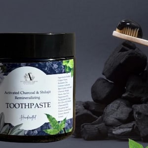 Può includere: Un barattolo di dentifricio remineralizzante al carbone attivo e shilajit con coperchio nero. Uno spazzolino di legno con dentifricio nero sulle setole poggia su un mucchio di carbone nero. Il barattolo ha un'etichetta bianca con testo e decorazioni floreali.