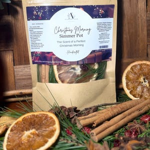 Może przedstawiać: Papierowa torba typu kraft z napisem "Christmas Morning Simmer Pot" z tekstem i okrągłym logo. Torba zawiera suszone plasterki pomarańczy, laski cynamonu i igły sosnowe. Widoczne są inne składniki.