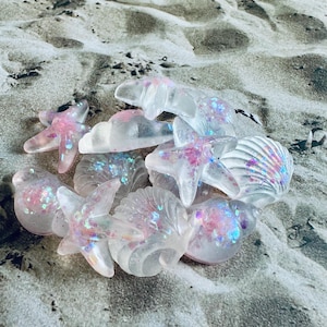 以下が含まれることがあります： ピンクと虹色のグリッターが付いた透明なレジン製の貝殻とヒトデのコレクション。貝殻とヒトデは砂浜の上に置かれています。