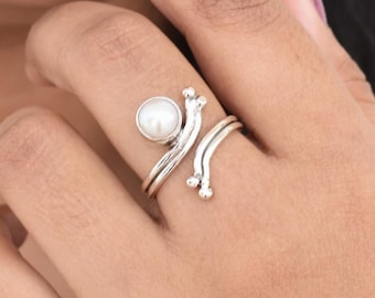 May include: A silver ring featuring a white pearl and a swirling design. The ring is worn on a person's finger.