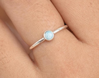 May include: A silver ring with a small, round, light blue gemstone. The ring is on a finger, and the hand is in front of a blurred background. The ring's band is thin and simple.