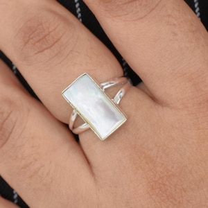 May include: A silver ring with a rectangular, iridescent mother-of-pearl stone. The ring is on a hand with pink nail polish, set against a black and white striped background. The ring's design features a split band.