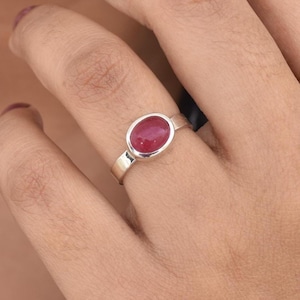 May include: A silver ring with a red oval gemstone. The ring is on a person's finger.