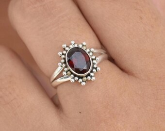 May include: Oval-shaped, dark red gemstone ring with a silver band. The gemstone is surrounded by a halo of small, beaded accents. The ring is on a finger, showcasing the jewelry's design and craftsmanship.