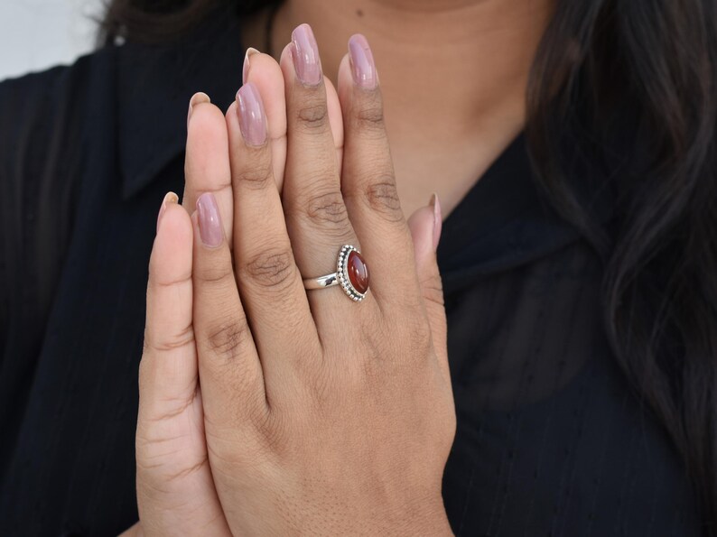 May include: A silver ring with a red oval gemstone set in a beaded bezel. The ring is on a person's finger.