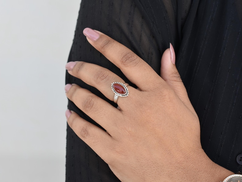 May include: A silver ring with a red carnelian gemstone set in a beaded bezel. The ring is on a person's finger.