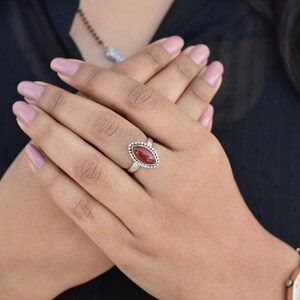 May include: A silver ring with a red oval gemstone set in a beaded bezel. The ring is on a person's finger.