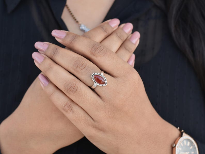 May include: A silver ring with a red oval gemstone set in a beaded bezel. The ring is on a person's finger.