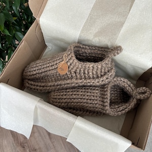 Könnte beinhalten: Ein Paar grob gestrickte braune Hausschuhe mit Rippenstruktur, versehen mit einem kleinen runden Anhänger, der mit einer Schnur befestigt ist und die Aufschrift "made with love" sowie ein Herzsymbol trägt, präsentiert in einem mit weißem Seidenpapier ausgelegten Karton.