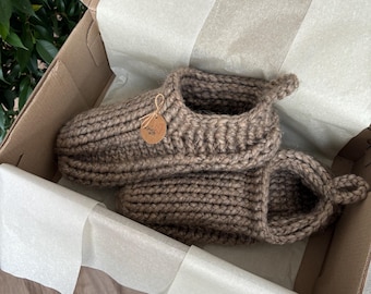 May include: A pair of chunky, hand-knitted brown slippers with a ribbed texture, featuring a small round tag tied with twine that reads "made with love" and a heart symbol, presented inside a cardboard box lined with white tissue paper.