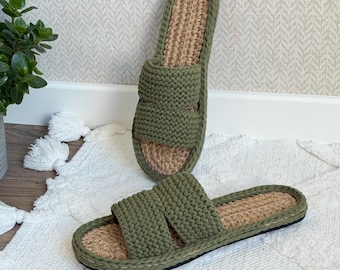 May include: Two olive green knitted slide sandals with a jute footbed are displayed on a white rug. The sandals have a thick, textured strap across the top and a black sole. One sandal is upright, and the other is lying flat.