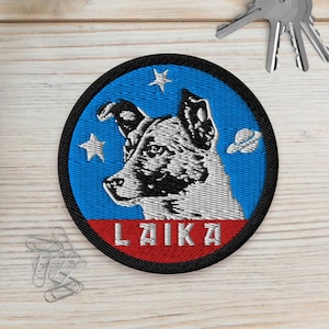 May include: Round embroidered patch with a dog portrait, stars, and a planet on a blue backdrop, with the name "LAIKA" in red. The patch has a black border. Two small potted succulents and keys are also visible.