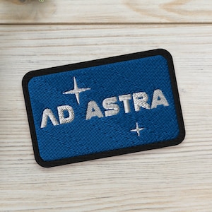 Könnte beinhalten: Ein blauer gestickter Aufnäher mit weißen Nähten, auf dem "AD ASTRA" mit zwei weißen Sternen auf jeder Seite des Textes steht. Der Aufnäher befindet sich auf einer hellen Holzoberfläche mit zwei kleinen Sukkulenten in Töpfen.