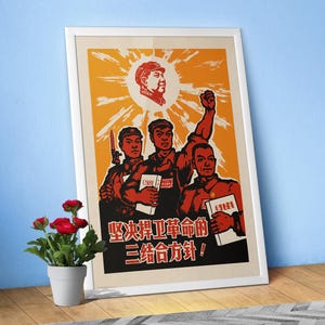May include: A framed poster featuring a stylized orange and black graphic illustration of three figures holding books, with a portrait of a leader in a sunburst above. The text reads 坚决捍卫革命的三结合方针. The poster is displayed in a white frame.