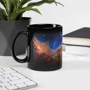 Puede incluir: Taza de café de cerámica negra con una imagen de nebulosa colorida impresa en ella. La nebulosa es una mezcla de naranja, rojo, azul y blanco.