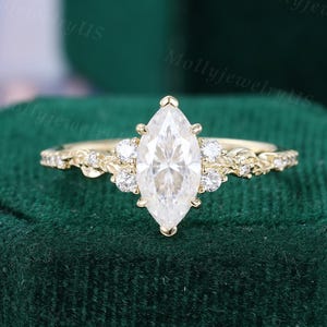 May include: A marquise-cut diamond engagement ring with a gold band. The ring features a large, central diamond and smaller accent stones. The band has delicate floral detailing. The ring is displayed on a dark green velvet surface.