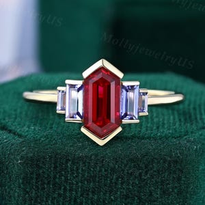 May include: A gold ring featuring a large, rectangular, deep red gemstone at the center, flanked by two smaller, rectangular, light blue gemstones on each side. The ring has a geometric design and is set against a dark green background.